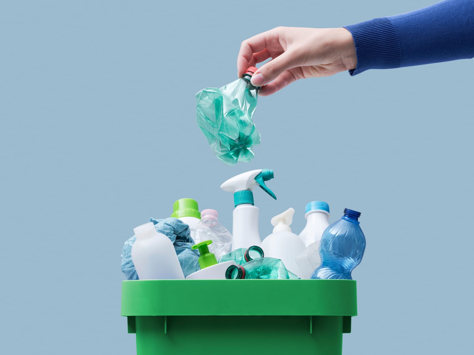 A hand drops a plastic bag into a green collection bin full of plastic bottles