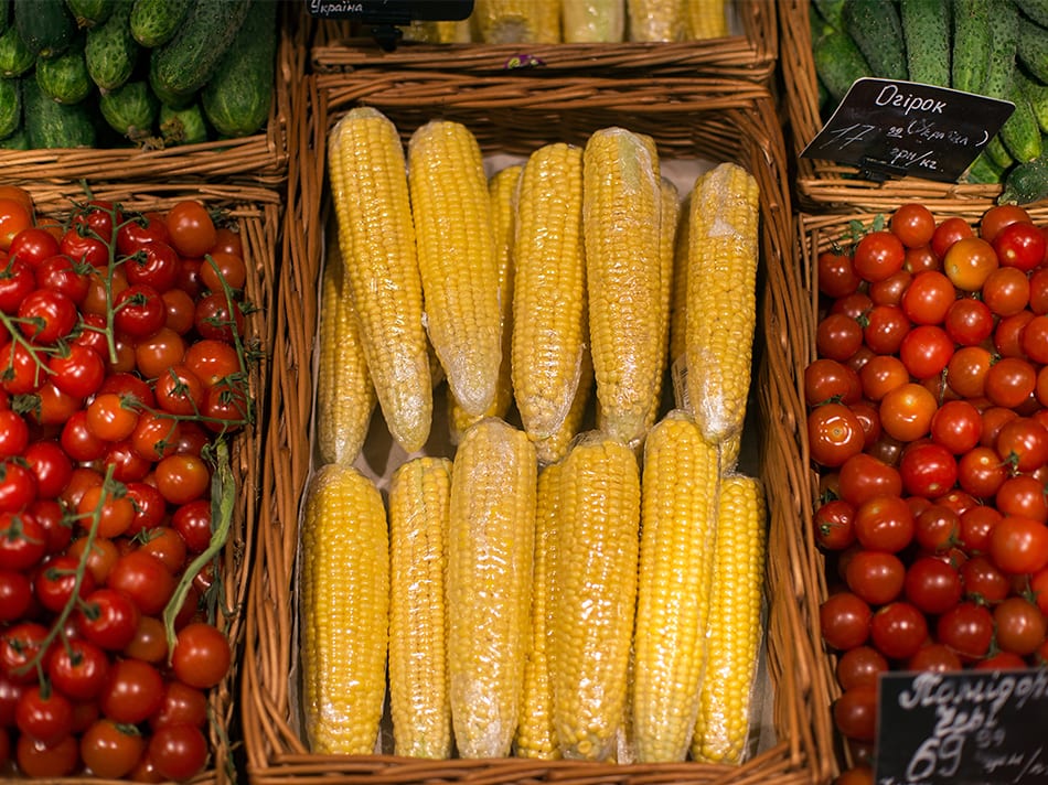 Film-wrapped corn cobs in a basket, with cherry tomatoes and cucumbers at a market stall