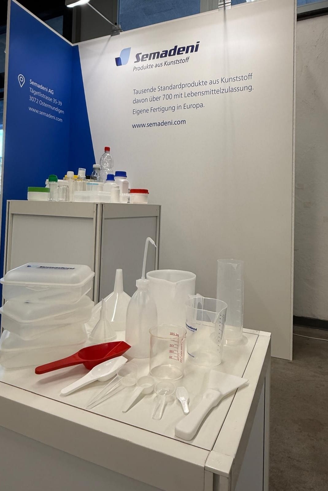 Ausstellungstheke am Semadeni-Stand mit Lebensmitteldosen, Trichtern, Spritzflasche, Messbechern, Messzylinder und Dosierlöffeln aus Kunststoff, dahinter die Standwand mit Semadeni-Logo