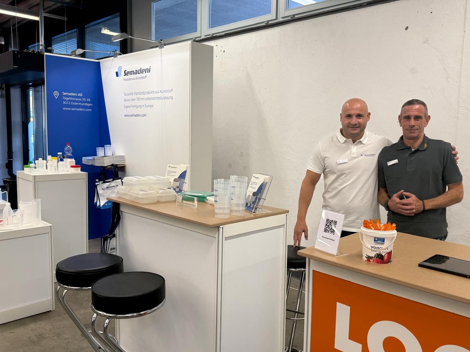Zwei Mitarbeiter am Semadeni-Stand E195 an der NovaFood 2026 in Luzern, einer im Semadeni-Poloshirt, einer im Poloshirt von Logo-Plastic; daneben Theken mit Lebensmitteldosen, Messbechern und Flaschen aus Kunststoff