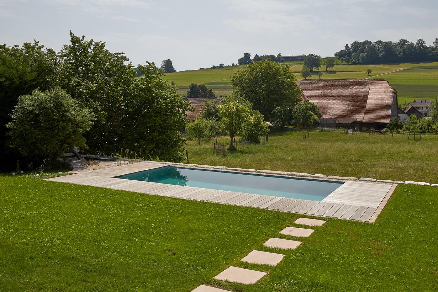 Rechteckiger Badeteich in der Wiese, mit Holzdeck und Trittplatten, dahinter das Bauernhaus