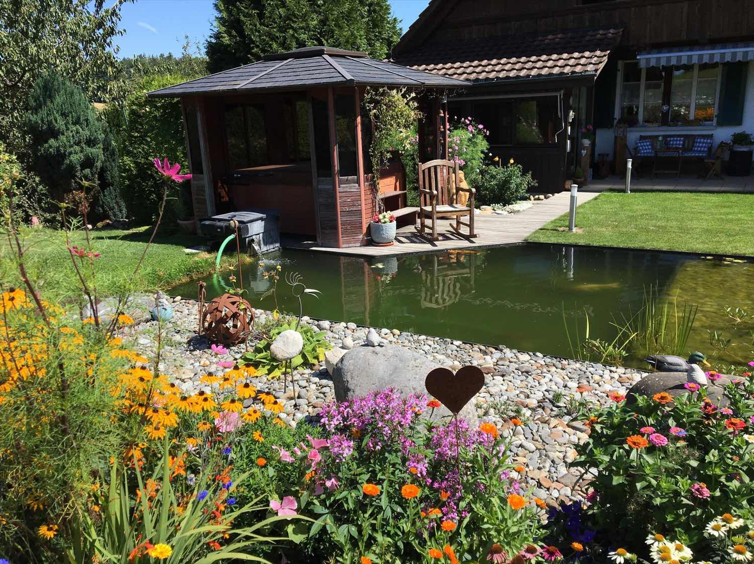View across the pond towards the covered seating area, the bank planted with perennials and stones