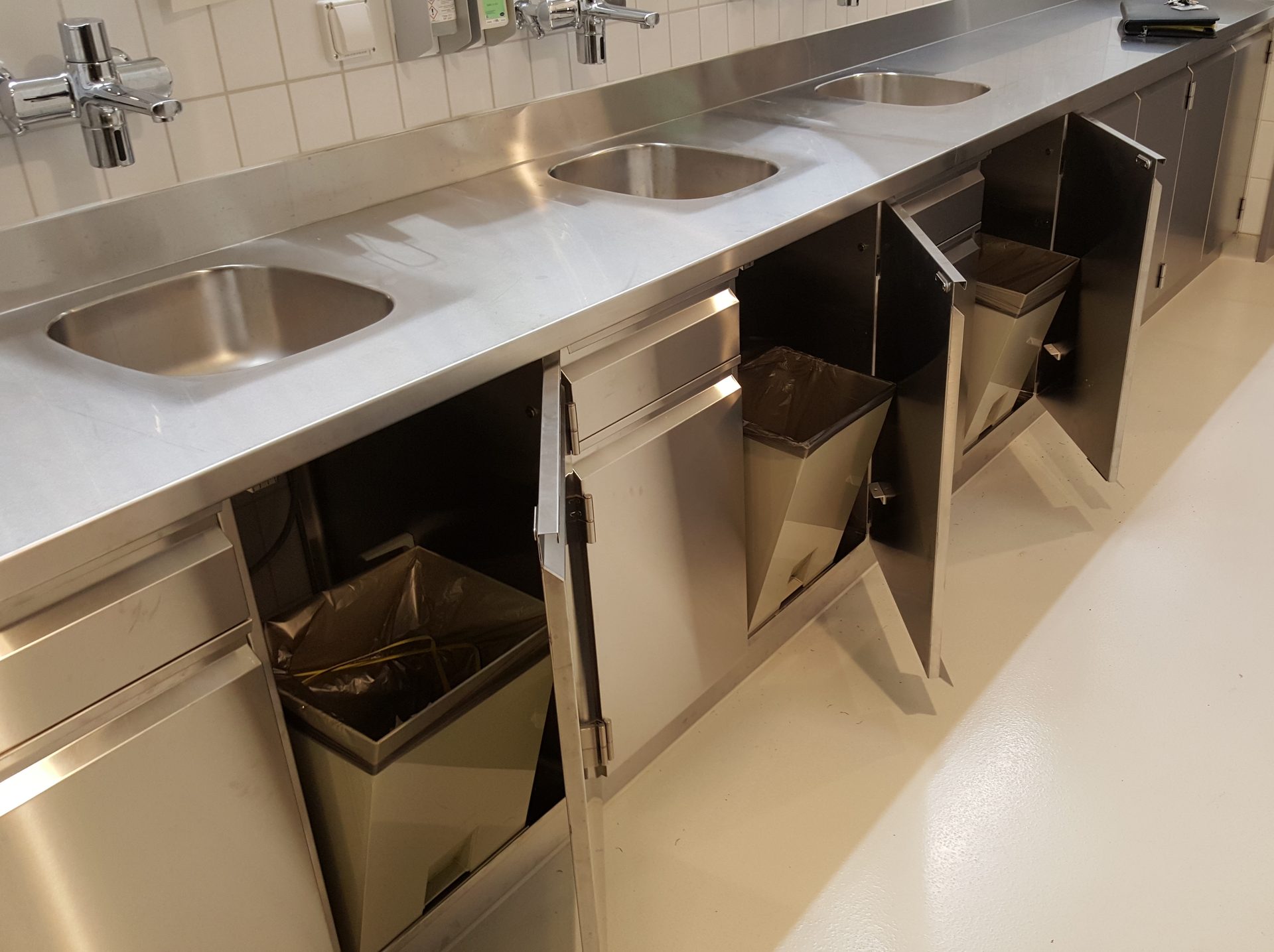 Grossküche mit Edelstahl-Spülen und ausgeschwenkten Abfallbehältern unter der Arbeitsfläche