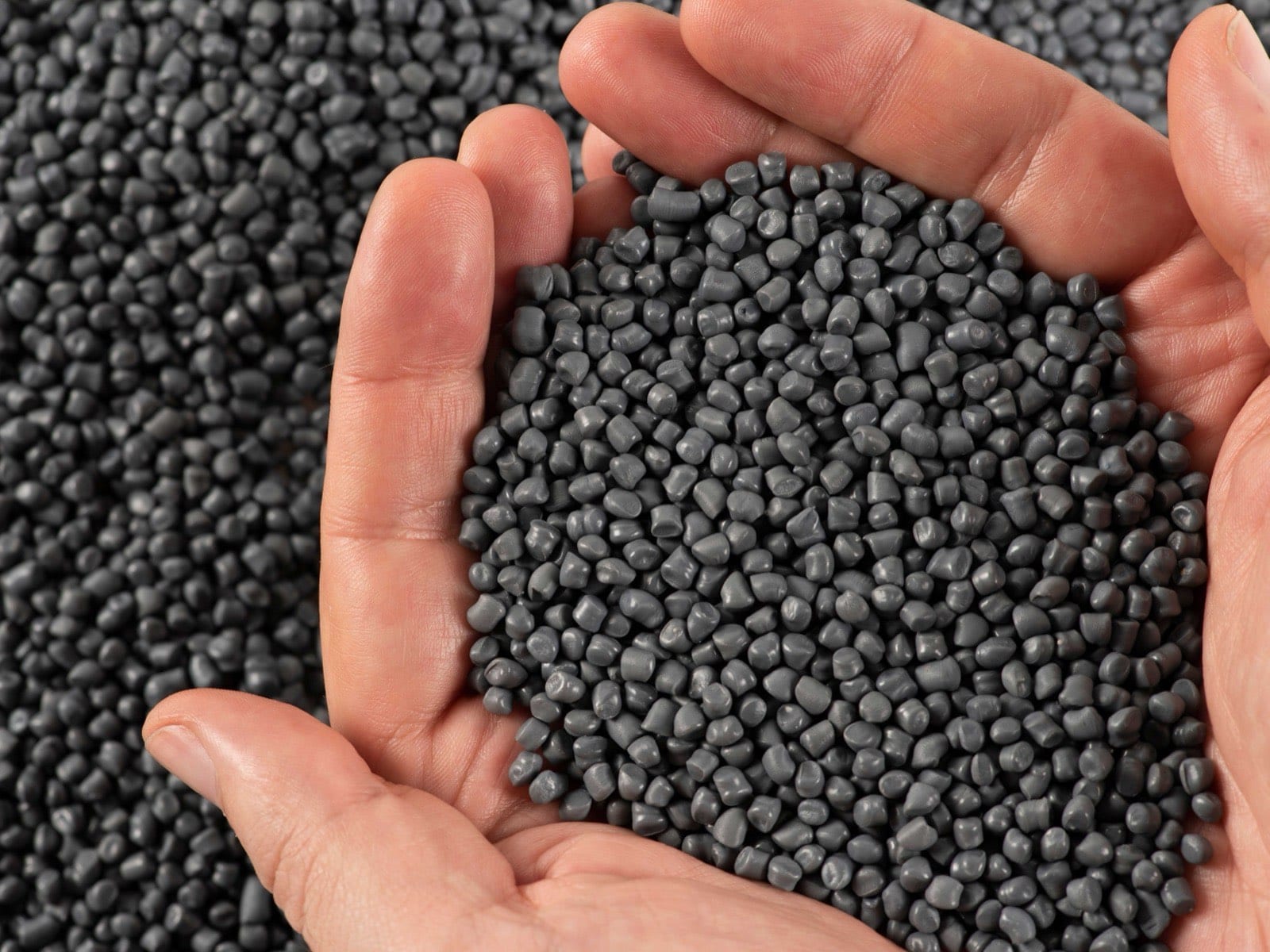 Hands holding grey plastic granulate above a heap of recyclate
